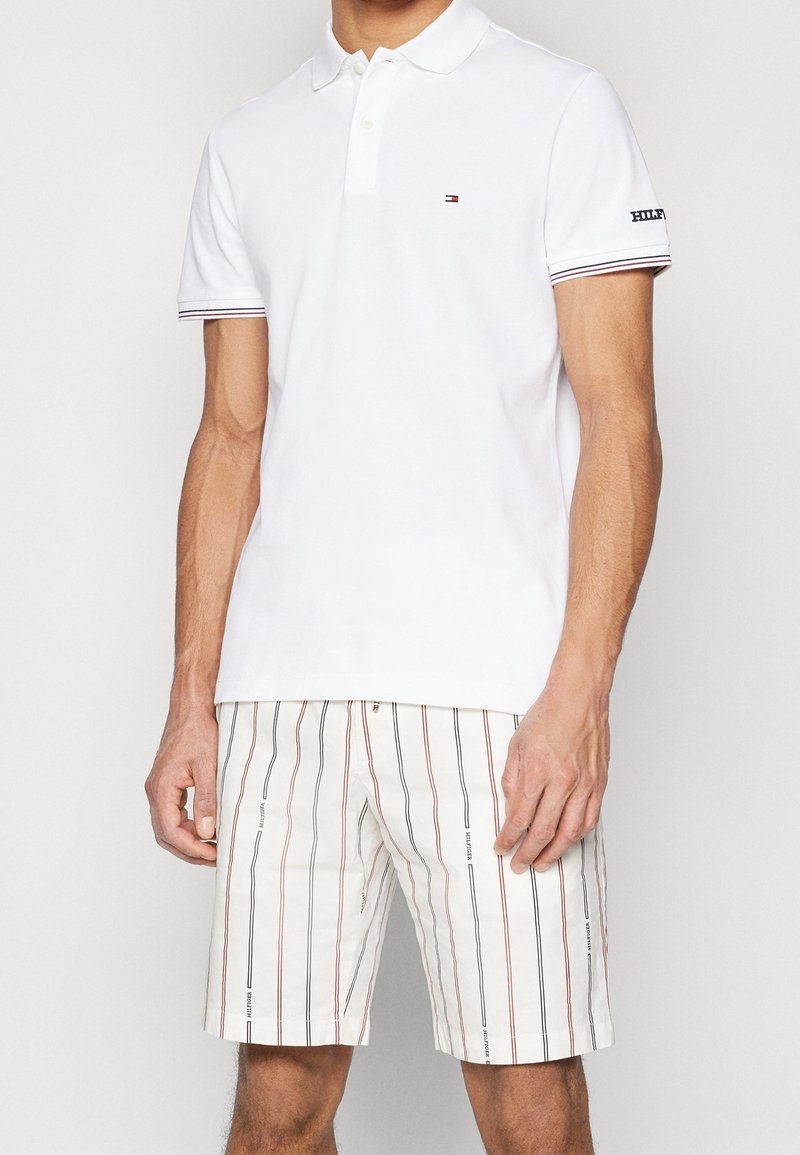 Homme portant un polo blanc à manches courtes et un short blanc avec des rayures verticales rouges et noires ainsi que des détails textuels.