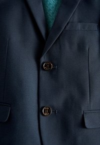 Blazer bleu foncé texturé avec deux boutons noirs, poches à rabat, et une cravate verte à motifs visible à travers le revers.