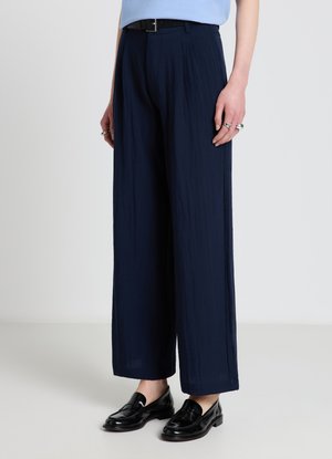 Persona che indossa pantaloni ampi blu navy, cintura nera, mocassini in pelle neri e una maglietta azzurro chiaro infilata, in piedi davanti a uno sfondo semplice.