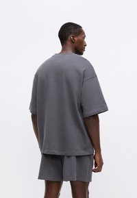 Hanorac gri cu mâneci scurte și croială lejeră, purtat împreună cu pantaloni scurți asortați. Textură netedă, design minimalist și decolteu rotund.
