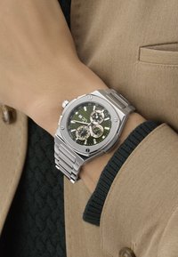 Hõbe roostevabast terasest kell rohelise kellapinnaga, kroonograafi funktsioonidega ja tekstuurse rihmaga. Märkimisväärsed detailid hõlmavad mitmeid alampaneele ja kuupäeva näidikut.