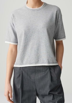 Persona con un suéter de manga corta color gris claro con ribete blanco y pantalones de rayas finas en gris oscuro, de pie con una mano en el bolsillo.