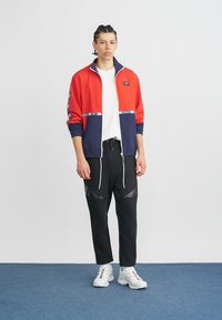 Junge Person steht in einer roten und marineblauen Jacke mit Reißverschluss, einem weißen T-Shirt, schwarzen Hosen mit Kordelzug und weißen Sneakers auf einem blauen Teppich.