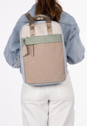 Frau mit einem Color-Block-Rucksack in Beige, Hellgrün und Grau mit goldenen Griffen, einer hellen Jeansjacke und weißen Hosen, von hinten gezeigt.