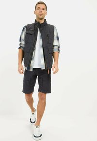 Marineblauer Weste mit mehreren Taschen über einem karierten Hemd, kombiniert mit dunkelgrauen Cargo-Shorts und weißen Sneakers mit marineblauen Akzenten.