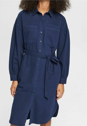 Kobieta ubrana w granatową sukienkę koszulową z długim rękawem, kieszeniami na piersi i paskiem zawiązanym w talii, na jednolitym tle.
