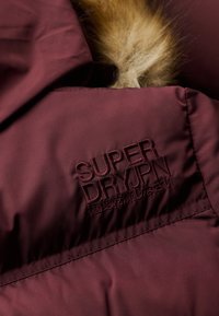 Bordeaux jack met een gladde textuur, voorzien van het reliëf "Superdry" logo en een bontaccent op de kraag.