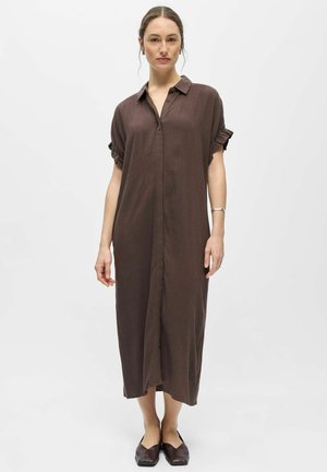 Kvinde med opsat hår iført en brun kjole til anklerne med korte flæseærmer og brune flade sko, stående mod en hvid baggrund.