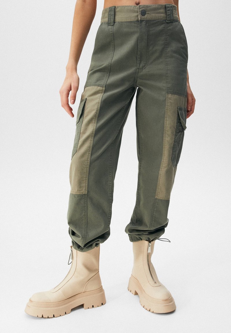 Olivové cargo kalhoty s lehčími panelovými akcenty, zúženými manžetami a bočními kapsami, doplněné béžovými kotníkovými botami na zip.