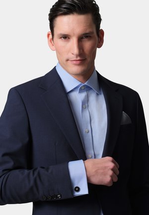 Blazer navy de tejido texturizado, camisa de vestir azul claro debajo, detalles de botones en los puños y un pañuelo en el bolsillo del pecho.