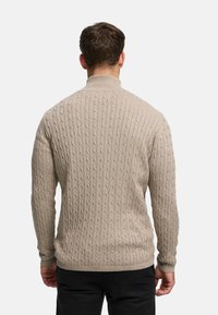 Beige kabelstrikket genser med høy hals, strukturert design og lange ermer, sett bakfra, kombinert med svarte bukser.