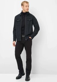 Giacca grigia scuro con zip e due tasche sul petto, indossata sopra un dolcevita nero. Abbinata a pantaloni neri e scarpe nere con lacci.