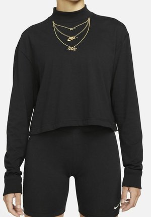 Femme portant un top court noir à manches longues, un short noir ajusté avec le logo Nike blanc et des colliers Nike dorés superposés.