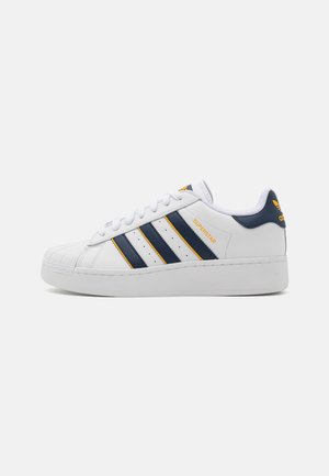 Weißer Adidas Superstar Sneaker mit marineblauen, gelb umrandeten Streifen und passendem Fersenlaschen-Logo, flacher Sohle und Shell-Toe-Design.