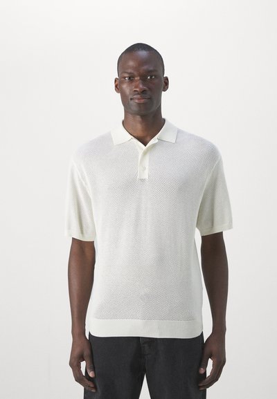 Polo shirt en maille beige clair avec col et manches courtes. Présente un motif texturé, une fermeture à boutons au cou et une coupe décontractée.