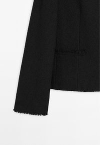Černý strukturovaný kabát s cropped designem, ozdobený třepícími se lemy na rukávech a jednou kapsou.