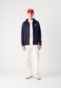 Marineblå zip-hoodie med to lommer, hvit t-skjorte under, lyse bukser og en rød caps med hvit tekst.
