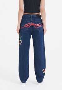 Blue denim jeans with a relaxed fit, featuring red embroidered "Ed Hardy" logo and colorful tattoo-style graphics on the legs.