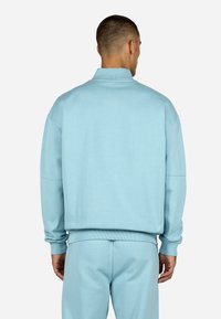 Gaiši zils džemperis ar augstu apkakli, brīvi pieguļošs un ar ribotām piedurknēm. Mīksta tekstūra, savienots ar atbilstošām biksēm. Aizmugures skats.