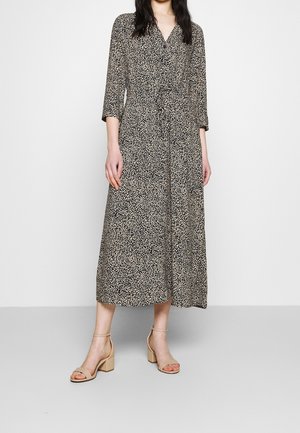 Kobieta w czarno-białej sukience midi z kwiatowym wzorem, z rękawami do łokcia, na beżowych sandałach na klocku, stojąca przy białej ścianie.