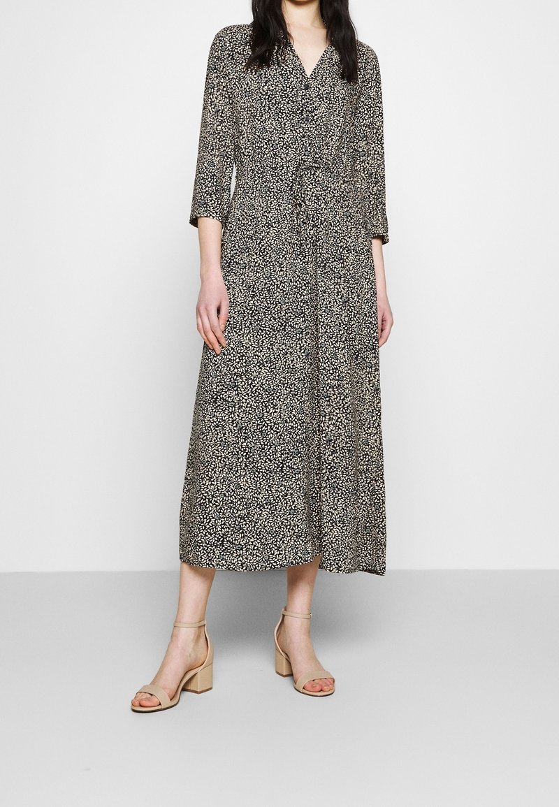 Femme portant une robe midi portefeuille à imprimé floral noir et blanc avec manches trois-quarts et sandales à talons blocs beiges, debout contre un mur blanc.