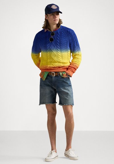 Striktrøje med gradient fra blå til gul til orange, med kabelstrikkemønster. Båret over en ternet skjorte, parret med denimshorts og hvide sneakers.