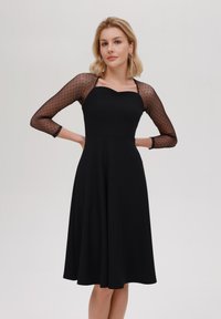 Černé šaty s přiléhavým korzetem a rozšířenou sukni. Mají průhledné rukávy s puntíkovaným vzorem. Hladká tkanina, elegantní design výstřihu.