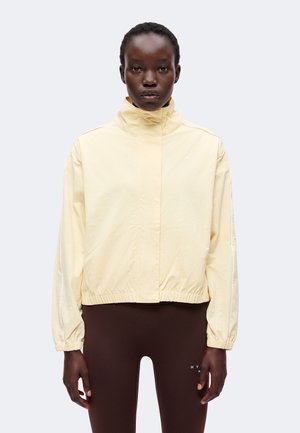 Person iført en lysegul jakke med høj krave og mørkebrune leggings, der står med fronten mod kameraet med et neutralt udtryk.