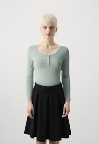 Langermet, lysegrønn topp med rund hals og knappedetalj, kombinert med et svart plissert skjørt med elastisk midje.
