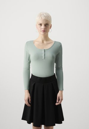 Langermet, lysegrønn topp med rund hals og knappedetalj, kombinert med et svart plissert skjørt med elastisk midje.