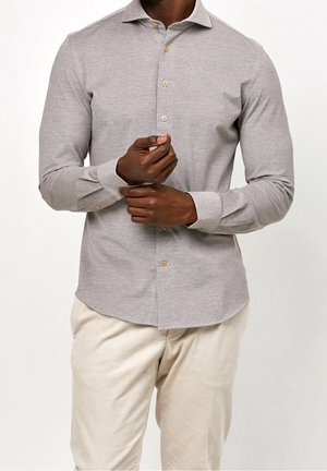 Homme ajustant la manche d'une chemise gris clair à manches longues boutonnée portée avec un pantalon beige sur fond blanc.