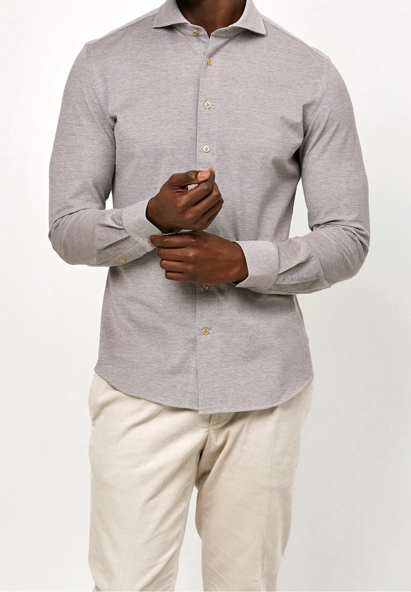 Homme ajustant la manche d'une chemise gris clair à manches longues boutonnée portée avec un pantalon beige sur fond blanc.