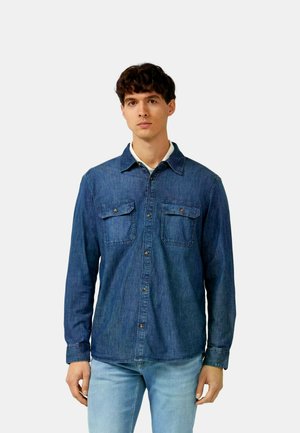 Junger Mann, der ein geknöpftes Jeanshemd mit Klappentaschen und hellblaue Jeans trägt, vor einem schlichten hellen Hintergrund stehend.