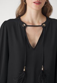 Black blouse with a V-neckline, gold grommets, and adjustable drawstrings featuring black feathered tassels. Smooth fabric with slight sheen.