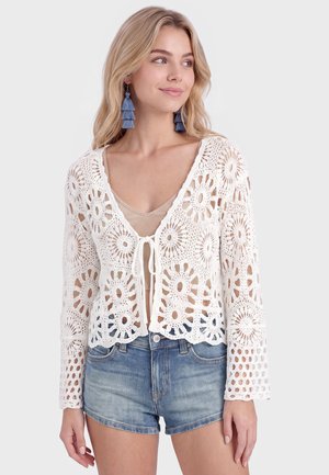 Lyshåret kvinde iført blå kvast øreringe, hvid hæklelangærmet top bundet foran, beige top under og denimshorts.