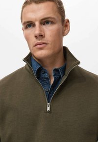 Sudadera verde oliva con cremallera y cuello alto, usada sobre una camisa de mezclilla azul. Presenta una cremallera plateada y una textura suave.