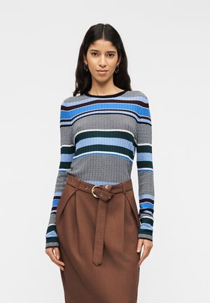 Žena má na sebe modré, čierne a biele pruhované tričko s dlhým rukávom a hnedú sukňu s vysokým pásom s opaskom v rovnakom odtieni.