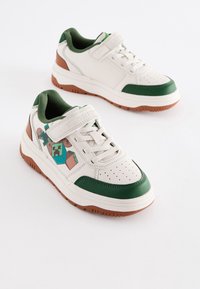 Baskets blanches avec des accents verts, dotées d'une bride à scratch, de détails perforés et d'un motif graphique sur le côté. Semelle en caoutchouc marron.