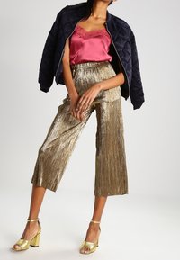 Mujer vestida con una chaqueta negra acolchada, un top de satén rosa, pantalones cortos plisados dorados y sandalias doradas de tacón alto.