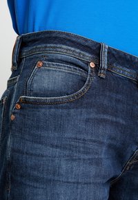 Jeans de denim azul oscuro con remaches y costuras de cobre, que presentan un diseño clásico de cinco bolsillos y una cintura de altura media. Textura suave.