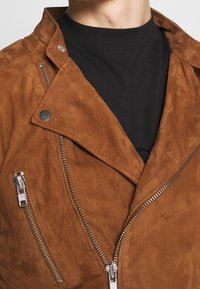 Giacca da moto in suede marrone con zip argentate, caratterizzata da un colletto a punta e un dettaglio con zip diagonale sul petto. Sotto, un dolcevita nero.
