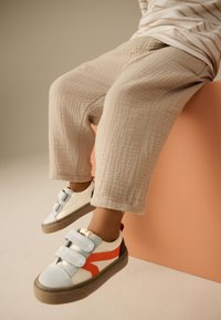 Baskets blanches et grises avec des accents orange et bleu, dotées de bandes Velcro, associées à un pantalon texturé beige clair.