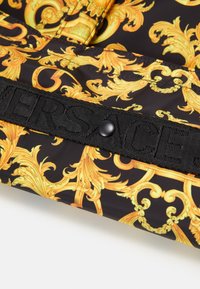 Gros plan sur un tissu à motifs baroques jaunes ornés sur fond noir, avec une sangle noire portant le texte en relief "VERSACE" et un bouton-pression.
