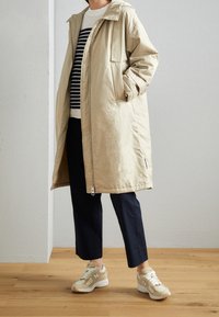 Abbigliamento esterno beige imbottito con cappuccio e zip frontale, con tasche laterali. Indossato sopra un maglione a righe, abbinato a pantaloni blu e sneakers.