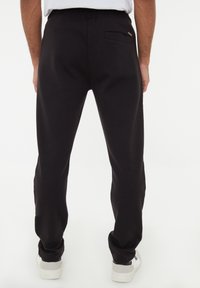 Schwarze Jogginghose mit einer lockeren Passform, elastischem Taillenbund und einer Gesäßtasche. Glatte Textur und minimalistisches Design, kombiniert mit weißen Sneakers.