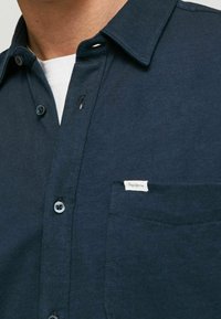 Camicia button-up blu navy in cotone, con colletto, tasca sul petto con etichetta e bottoni rotondi. Texture liscia.