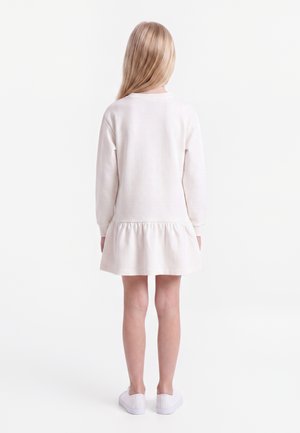 Młoda dziewczyna o długich blond włosach, ubrana w białą sukienkę z długim rękawem i białe trampki, stoi tyłem na gładkim białym tle.