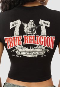 Czarny krótki t-shirt z dużym białym nadrukiem oraz czerwonym napisem „TRUE RELIGION”, przedstawiający postać Buddy z gitarą i banery z trasą koncertową.