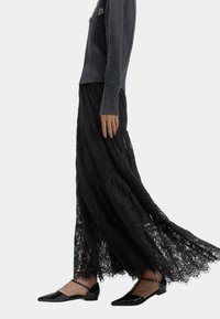 Svart maxikjol i spets med lager-på-lager-design, med vågig fåll; kombinerad med svarta lackplatta skor och en grå stickad tröja.