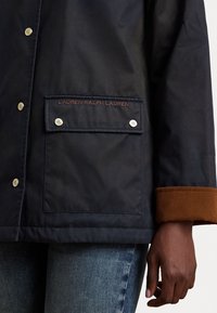Navy waterproof jacket with silver snaps, a side pocket, and brown ribbed cuffs. Features engraved brand detail on the pocket.